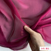 A hand is folding a piece of sheer magenta silk chiffon fabric.