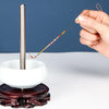 A hand is using a curved stainless steel beading needle to thread colorful beads onto a white ceramic tool.