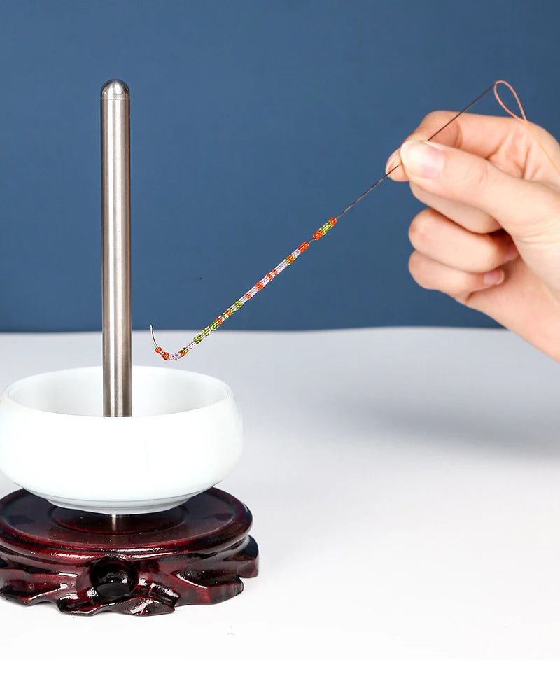 A hand is using a curved stainless steel beading needle to thread colorful beads onto a white ceramic tool.