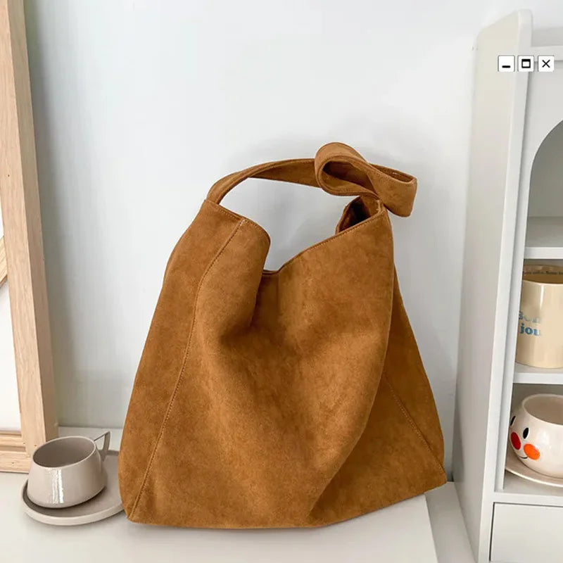 A tan suede tote shoulder bag is displayed on a white surface next to a light wooden frame and a white shelving unit.