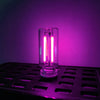 A glass cylindrical LED night light with a warm filament glow is placed on a dark metal tray against a purple background.