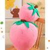 A pink strawberry plush throw pillow with green leaves is displayed on a wooden surface.