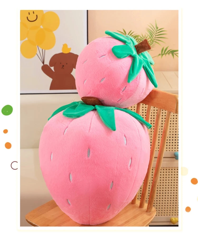 A pink strawberry plush throw pillow with green leaves is displayed on a wooden surface.