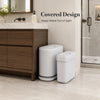 A white, rectangular trash can set with lids is placed on a light-colored floor next to a wooden cabinet and a glass shower e