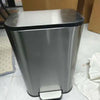 A stainless steel trash can with a lid and step pedal sitting on a light-colored floor.