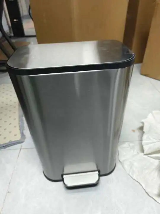 A stainless steel trash can with a lid and step pedal sitting on a light-colored floor.