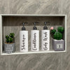 Three white refillable shampoo and lotion dispenser bottles with black pump tops displayed in a light gray shelf with green c