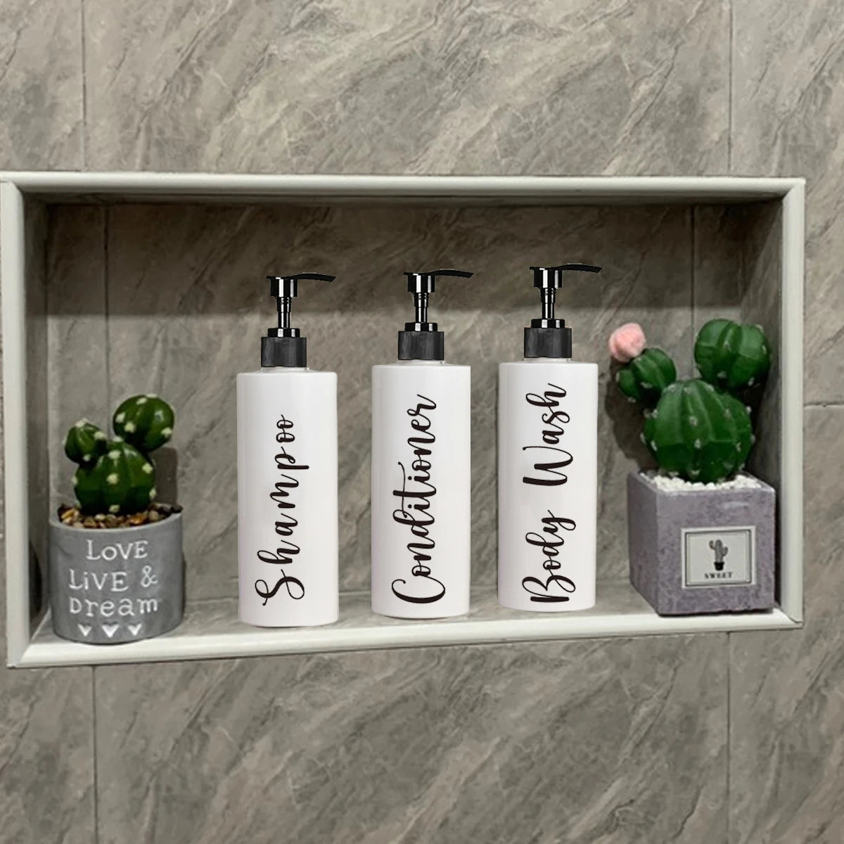 Three white refillable shampoo and lotion dispenser bottles with black pump tops displayed in a light gray shelf with green c