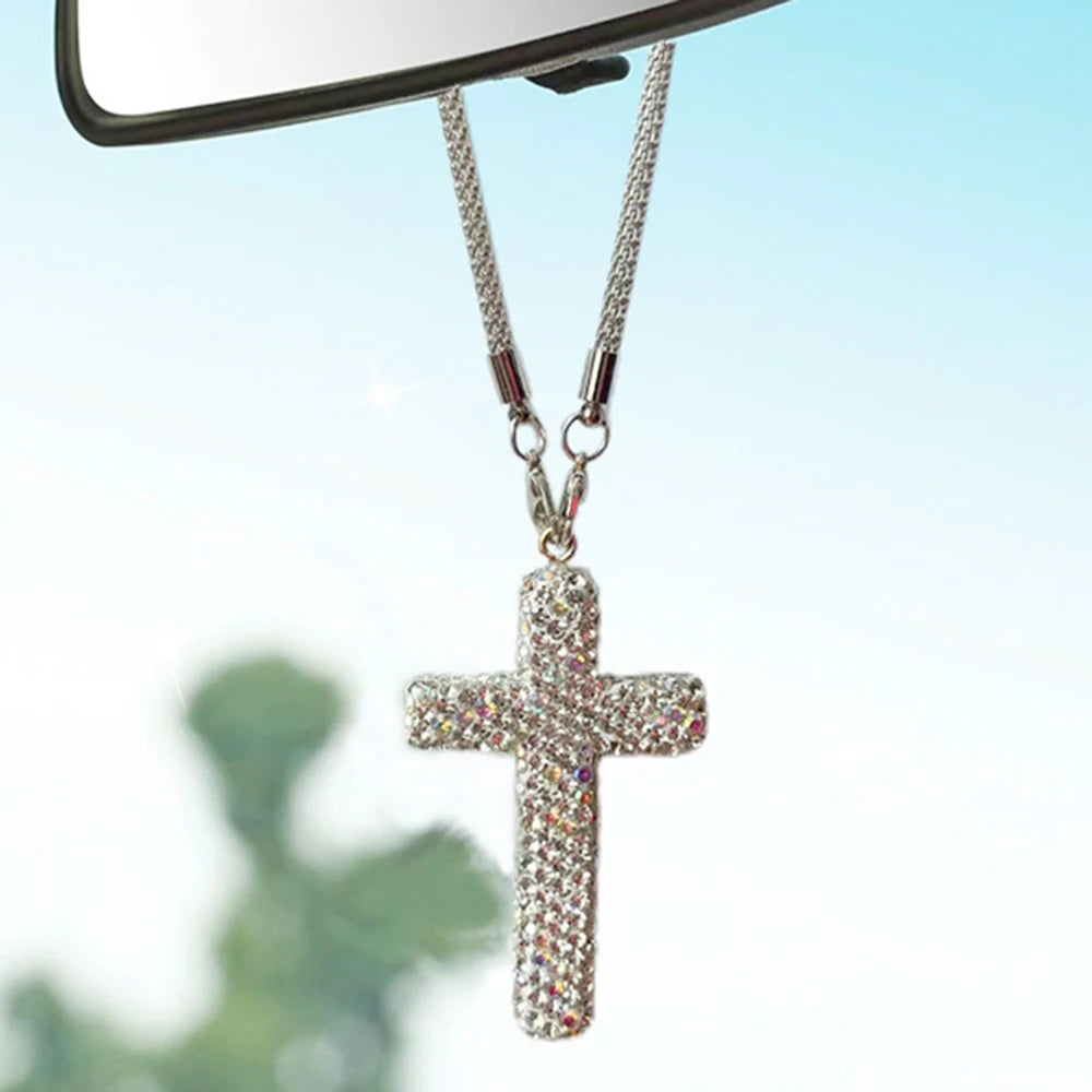 A silver stainless steel cross pendant adorned with rhinestones hanging from a chain, viewed against a blurred green backgrou
