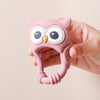 A hand holds a pink owl-shaped silicone teether and rattle for babies.