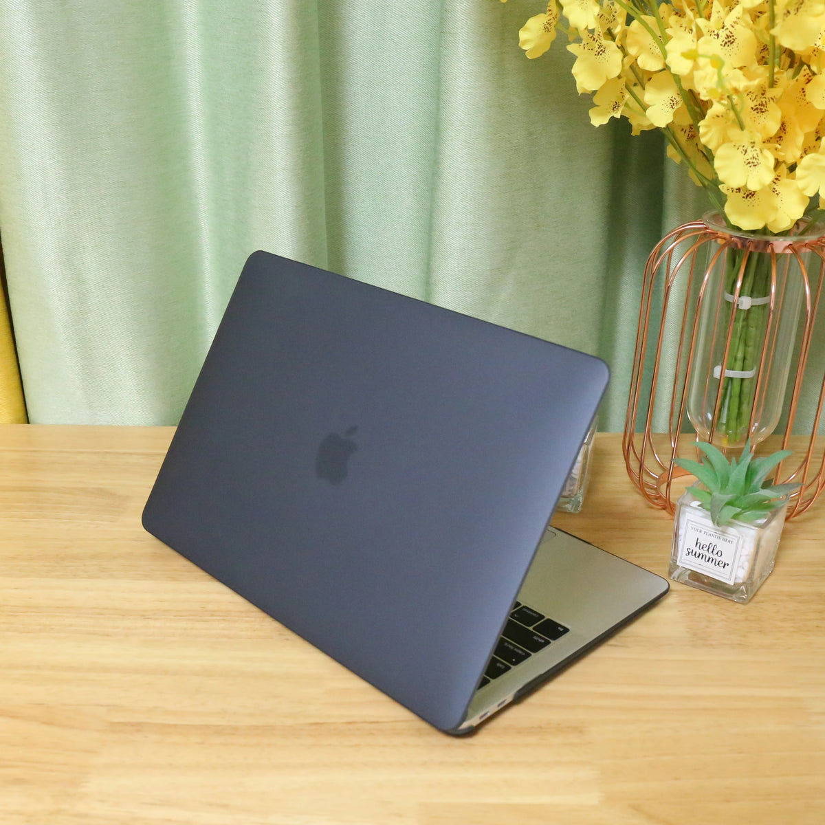 A dark gray MacBook is placed on a wooden table next to a glass vase filled with yellow flowers.