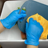 A hand wearing blue nitrile gloves is holding a dark gray plastic tool and an orange sponge in a white basin.