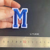 A hand holds a blue embroidered letter M patch, measured against a ruler.