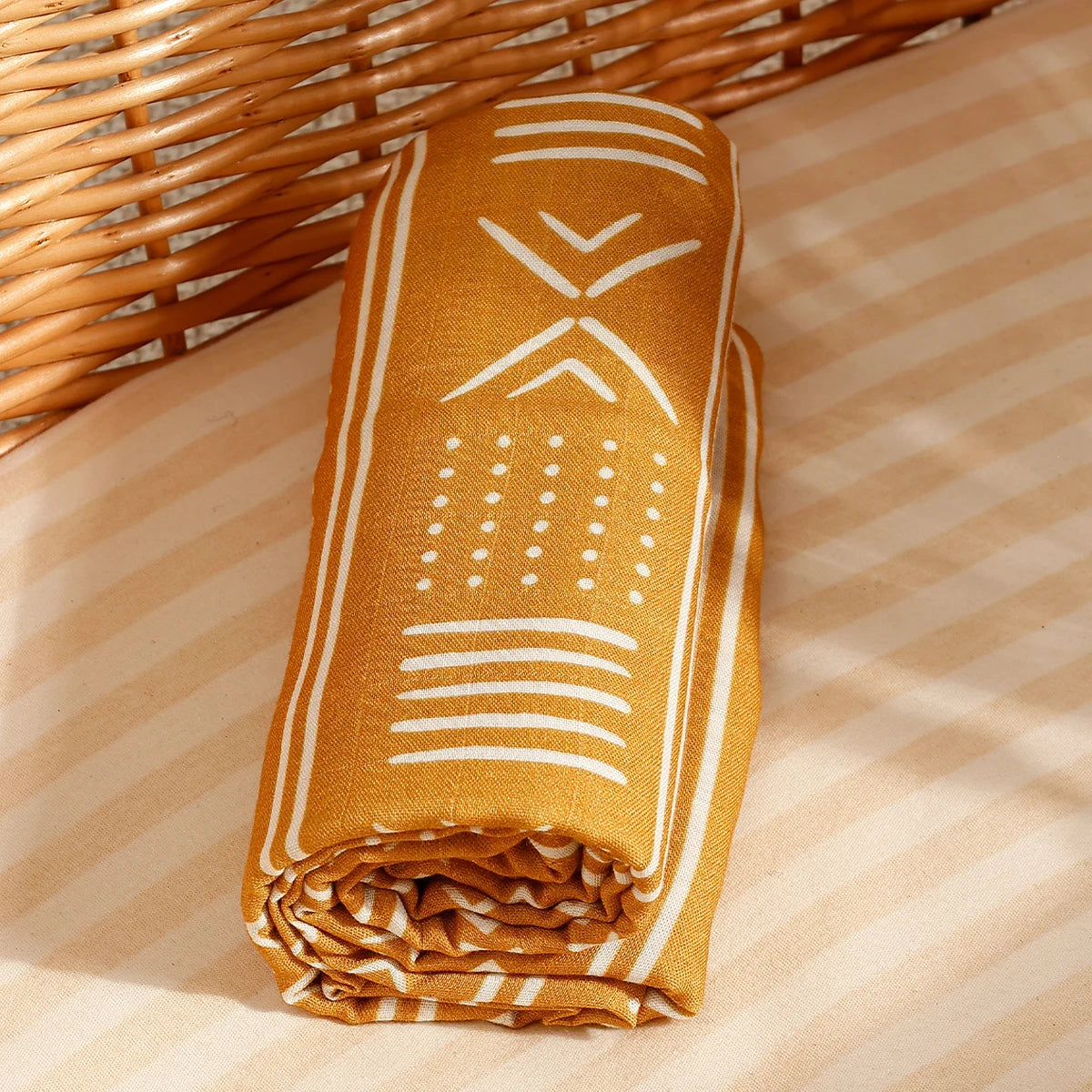 A rolled muslin baby swaddle blanket in mustard yellow with white geometric patterns resting on a light striped surface.