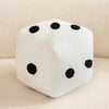 A white dice plush pillow with black dots, resting on a light-colored surface.