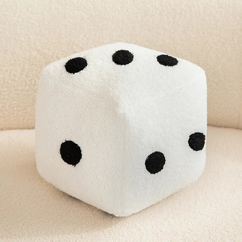 A white dice plush pillow with black dots, resting on a light-colored surface.