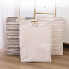 A beige striped foldable laundry basket with wooden handles standing on a wooden floor.