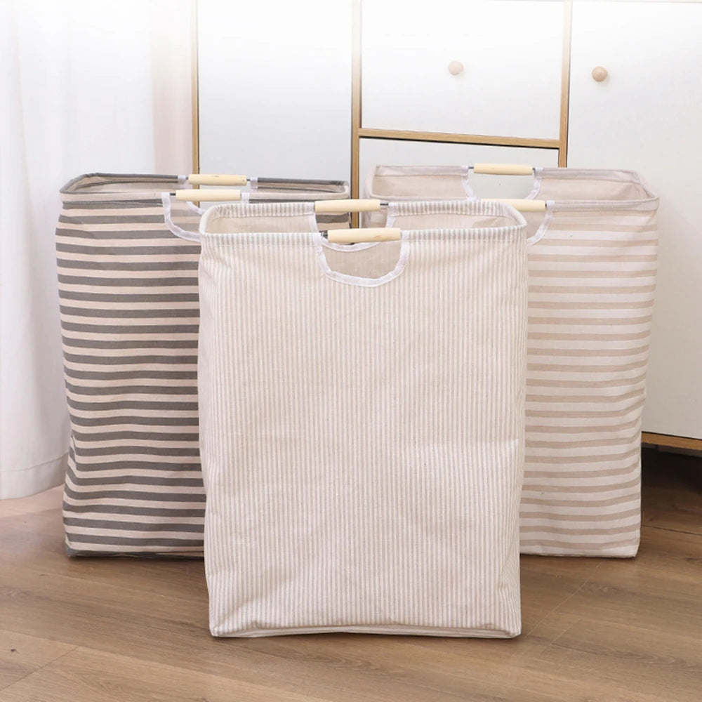 A beige striped foldable laundry basket with wooden handles standing on a wooden floor.