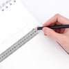 A hand holds a black pen against a stainless steel double-sided precision ruler on a white sheet of paper.