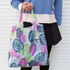A person holds a white and green floral patterned shopping tote bag against a white background.