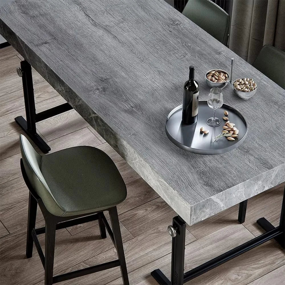 A modern dining table with a gray wood grain surface, featuring a dark bottle and nuts on a silver plate.