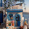 A charming seaside building facade featuring blue accents and a tree with purple blossoms, set against a clear blue sky.