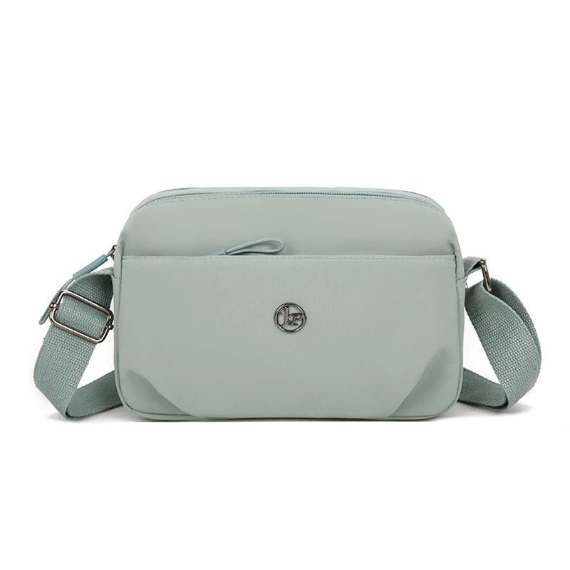 A light sage green nylon crossbody bag with a flap and adjustable strap is shown against a white background.