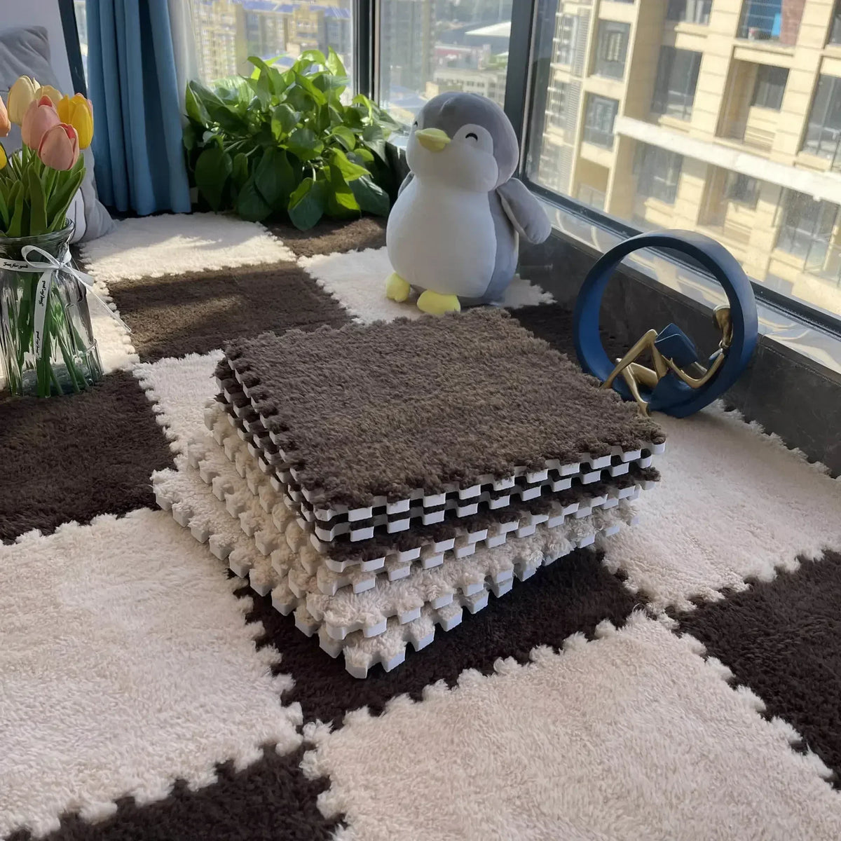 A stack of brown and white interlocking plush floor tiles with a gray stuffed animal on a light rug near a window.