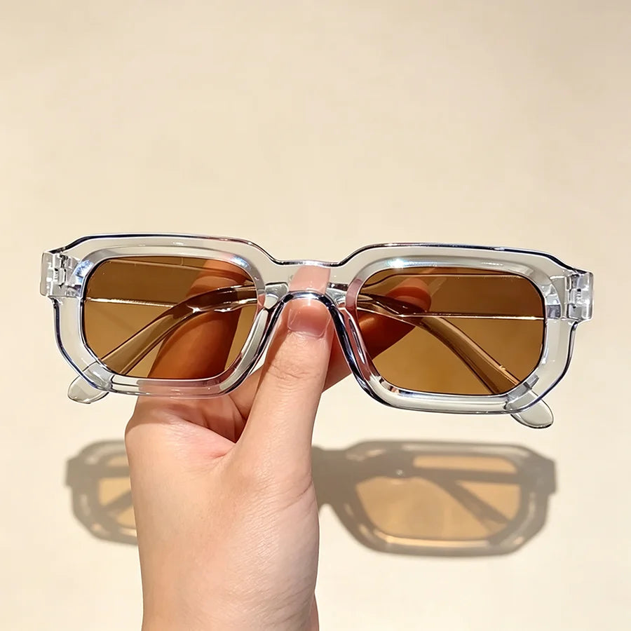 A hand holds a pair of vintage rectangle sunglasses with amber lenses against a light background.