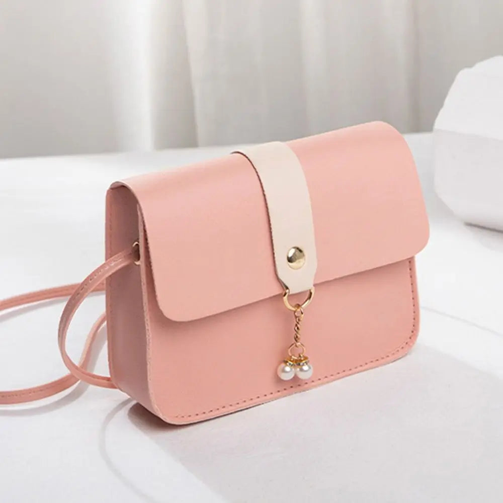 A light pink compact crossbody handbag with a beige adjustable strap and gold chain charm resting on a white surface.