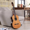 A wooden classical guitar is resting on a light gray sofa in a well-lit room.