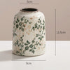 Weiße Keramikvase mit grünen und braunen Blumenmustern vor neutralem Hintergrund.