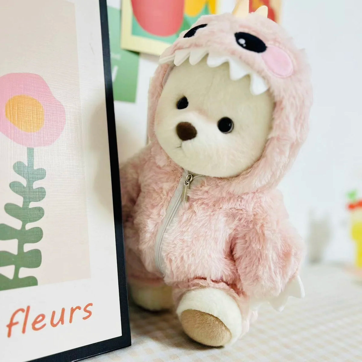 A pink fuzzy dinosaur jumpsuit outfit for a medium teddy bear doll displayed next to a floral illustration.