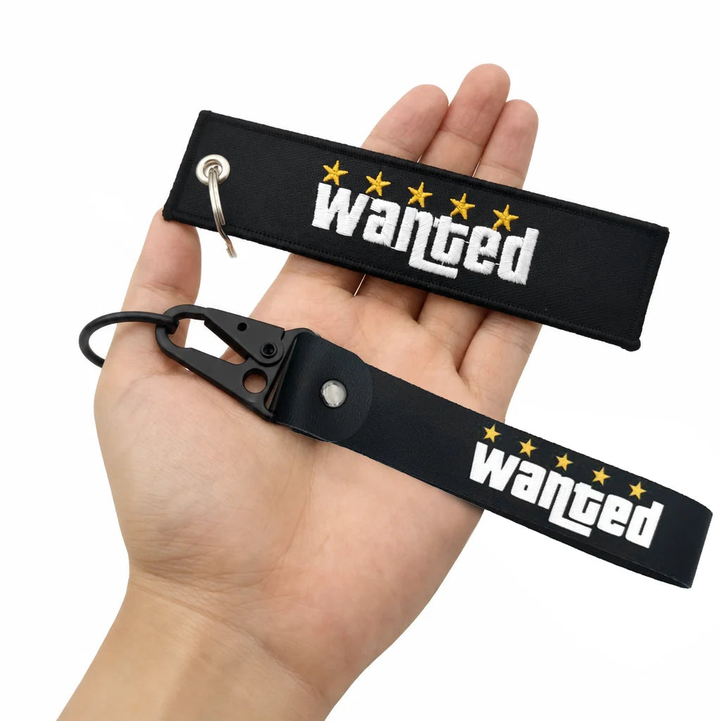 Black nylon keychain with embroidered wanted text and gold stars, held in a hand.