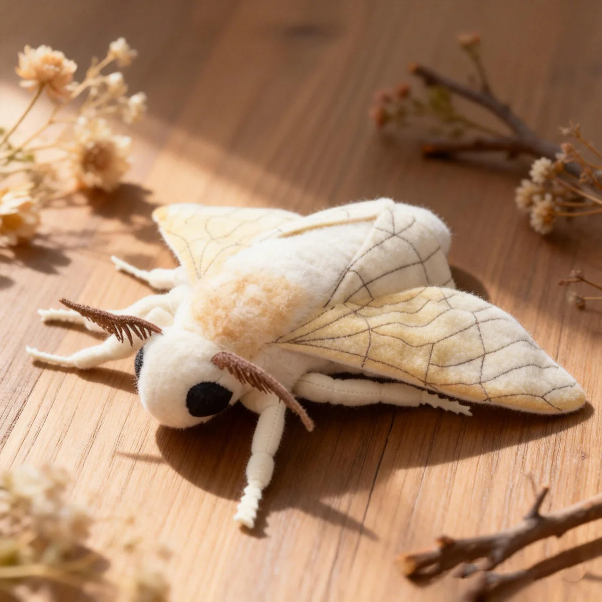 A white and cream Luna moth plush toy resting on a wooden surface with dried plant material.