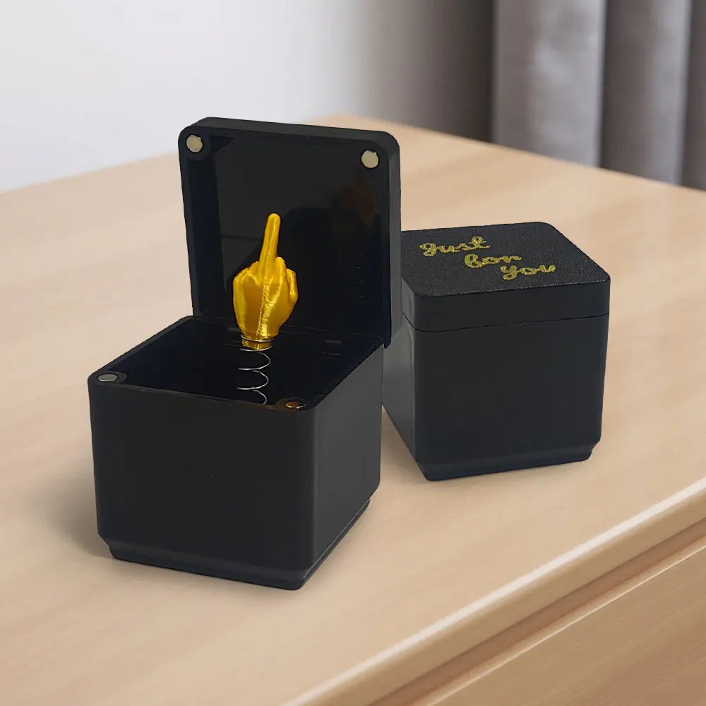 A black box with an open lid revealing a golden middle finger surprise inside