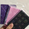 A hand holds a clear plastic package containing holographic heart-shaped card sleeves in various colors.