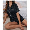 A model wearing a black satin pajama set with a button-down top and shorts, displayed in a studio setting.