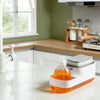 A white countertop soap dispenser with an orange liquid and a built-in sponge holder sits on a white counter.