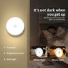 A white motion sensor LED night light illuminates a bedroom setting, demonstrating its portable and soft light features.
