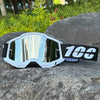 White motocross goggles with reflective lenses and a black strap resting on a gray rock.