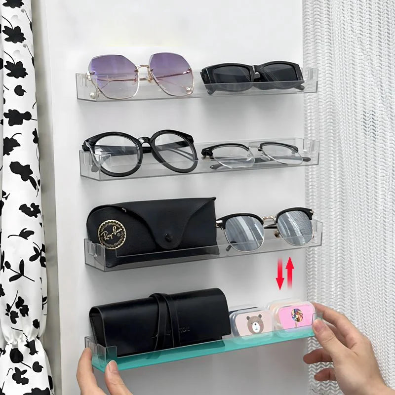A hand displays a clear acrylic wall-mounted display shelf holding various pairs of eyeglasses and sunglasses.