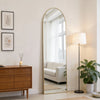 A full length oval wall mirror with a gold frame reflecting a bright room and window.