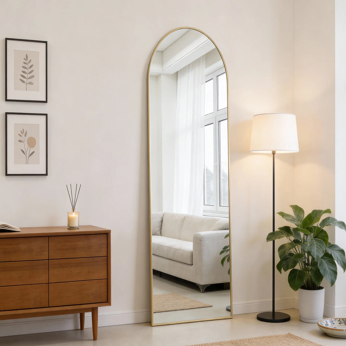A full length oval wall mirror with a gold frame reflecting a bright room and window.