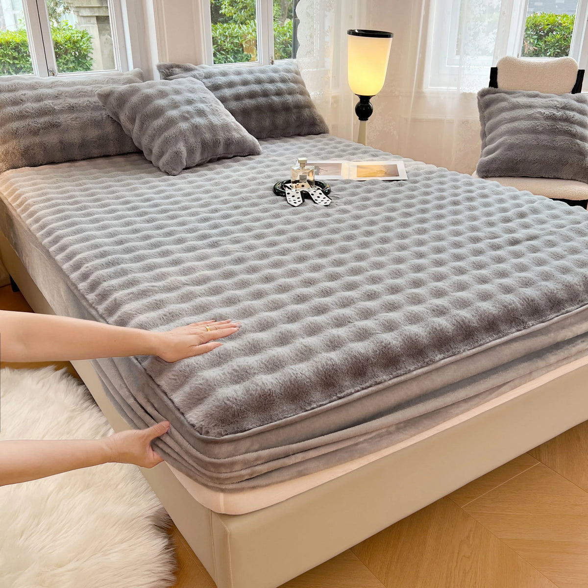 A gray luxury faux fur fitted sheet and pillowcase set is displayed on a bed, with a hand touching the fabric.