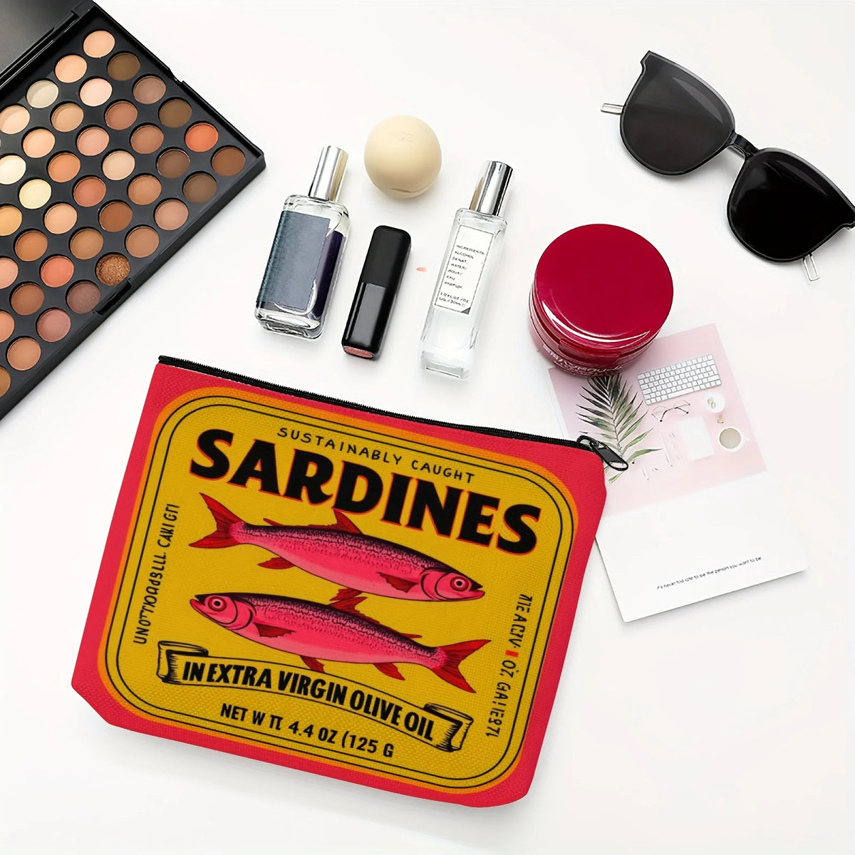 Pochette de maquillage rouge et jaune avec motif sardine, entourée de bouteilles en verre et de lunettes de soleil.