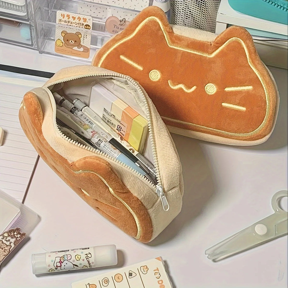 A caramel cat plush pencil case with a zipper is shown next to some stationery items.