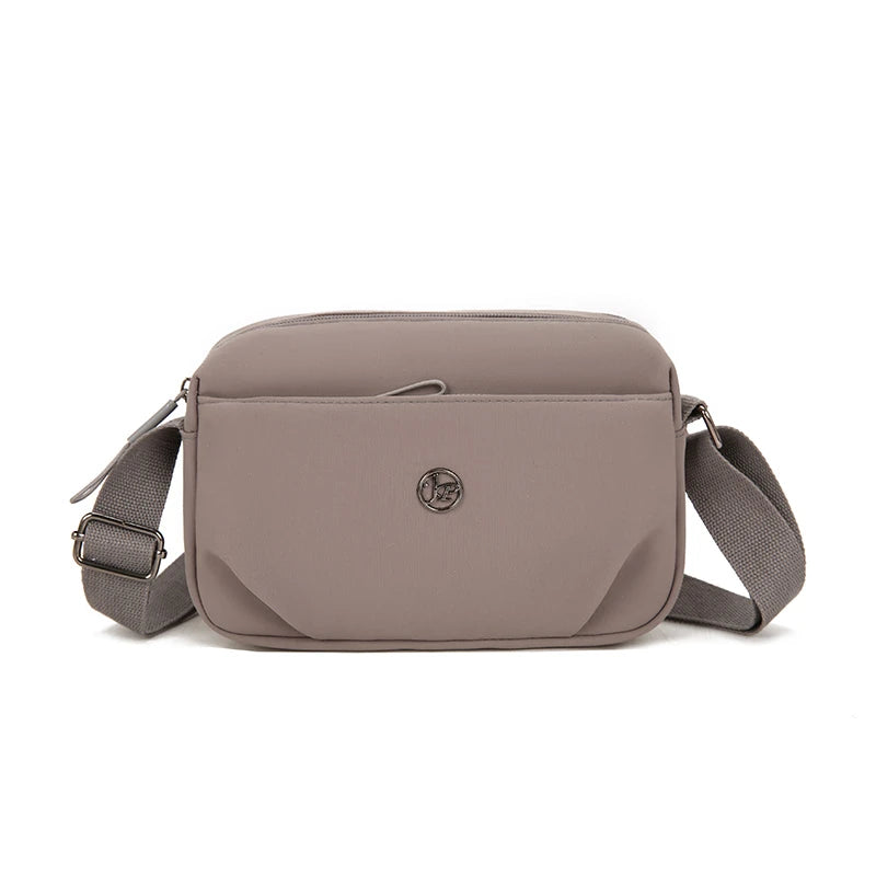 A light taupe nylon crossbody bag with a flap closure and adjustable strap is shown against a white background.