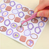 A hand uses a pink precision craft tool to pick up a piece of paper with various shaped stickers.