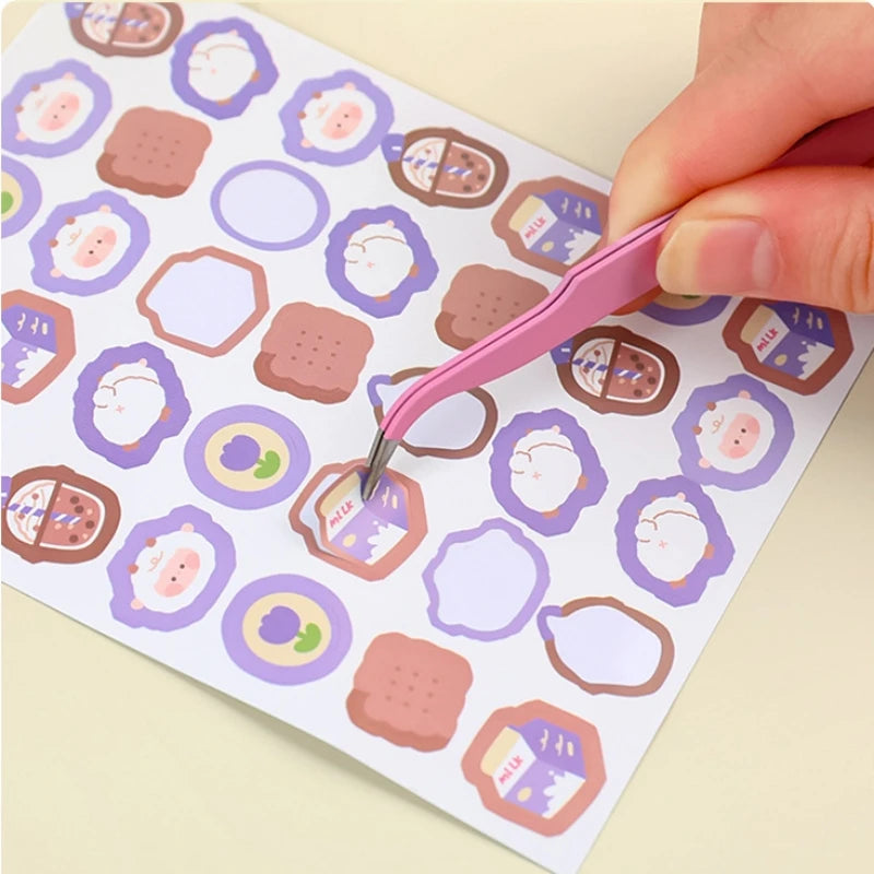 A hand uses a pink precision craft tool to pick up a piece of paper with various shaped stickers.